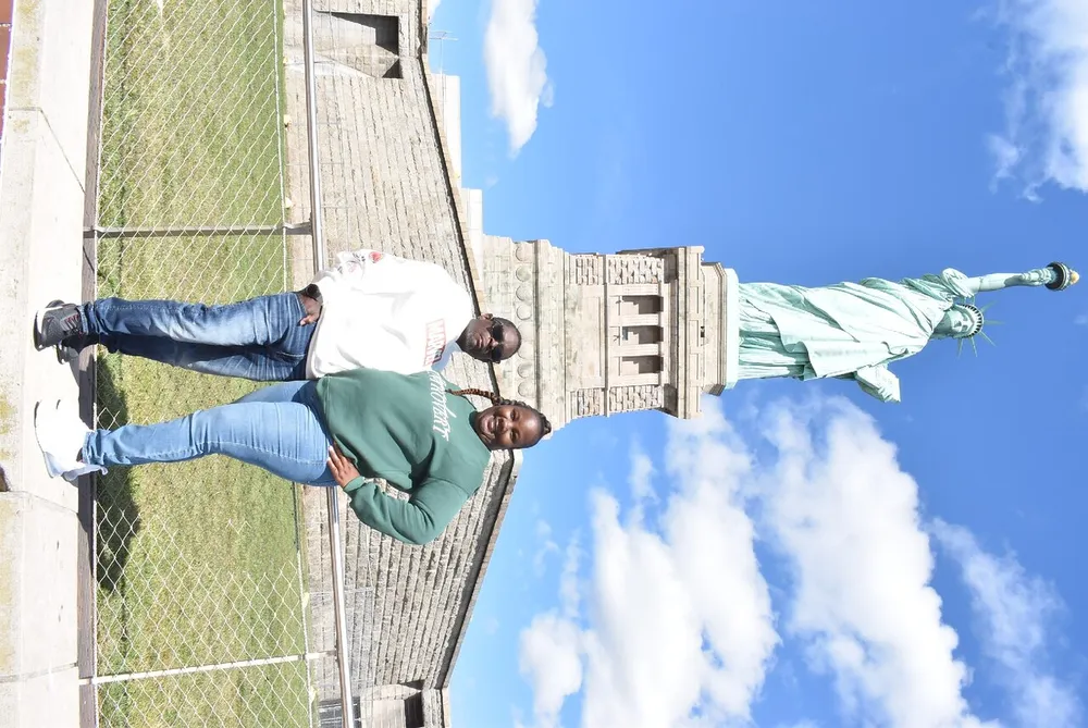 Two individuals are posing for a photograph in front of the Statue of Liberty on a sunny day with a clear blue sky in the background