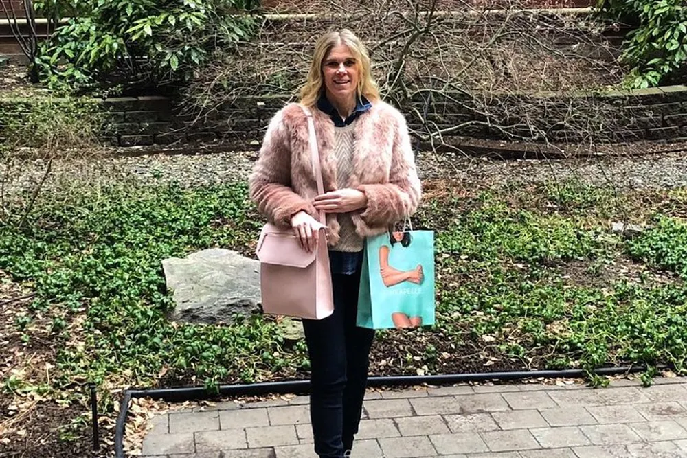 A woman in a pink furry jacket stands outside holding a light pink handbag and a shopping bag with greenery in the background