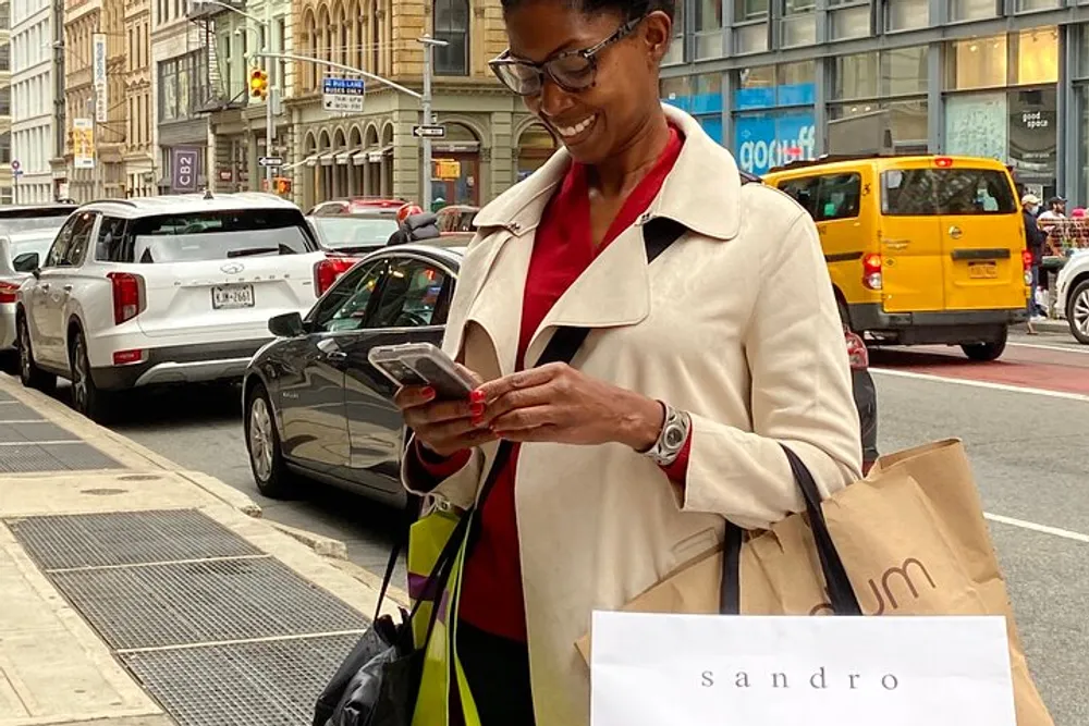 A smiling person is looking at their smartphone on a busy street corner holding shopping bags