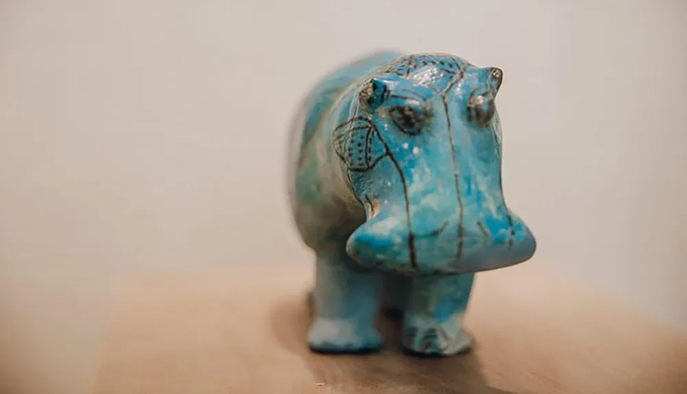 The image features a small blue faience sculpture of a hippopotamus with patterned details on a beige surface