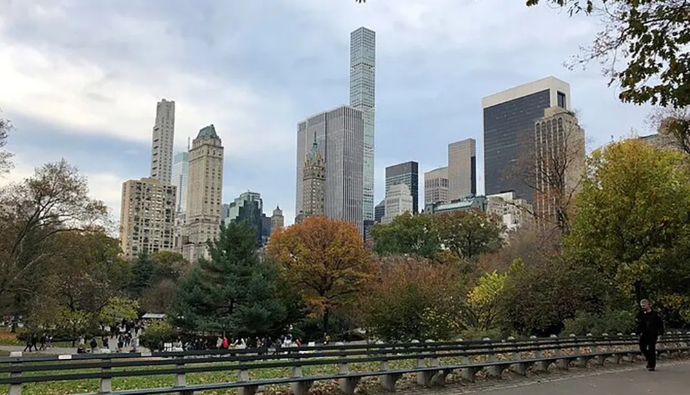 The image depicts a city park with people walking along paths surrounded by autumn-colored trees and a backdrop of urban high-rise buildings implying a blend of nature and city life