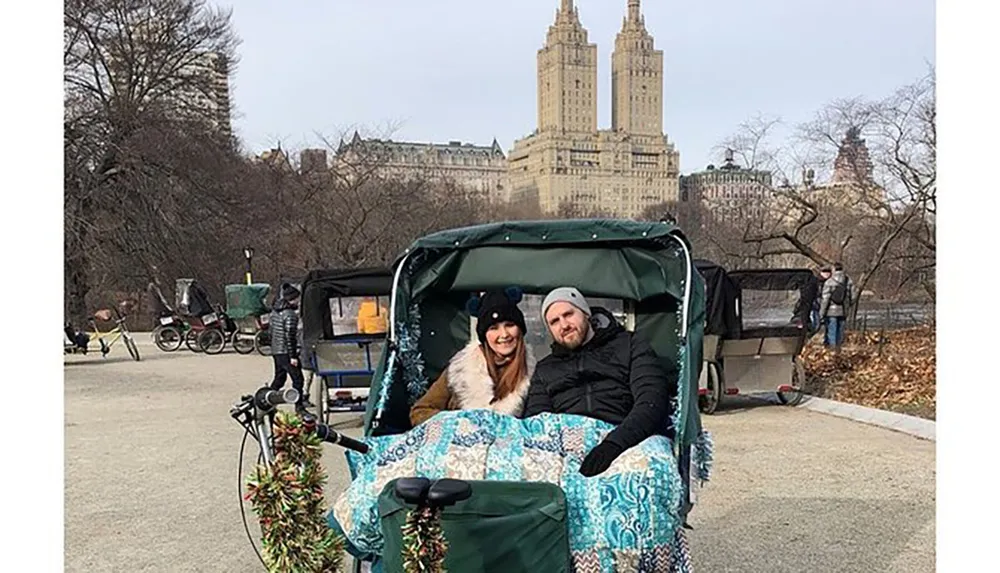 A smiling couple is bundled up under a blanket enjoying a pedicab ride in a park with leafless trees and a tall building in the background