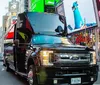 A black Ford truck with its hood open is parked on a busy street in Times Square amid bright digital billboards and passing traffic including a yellow cab
