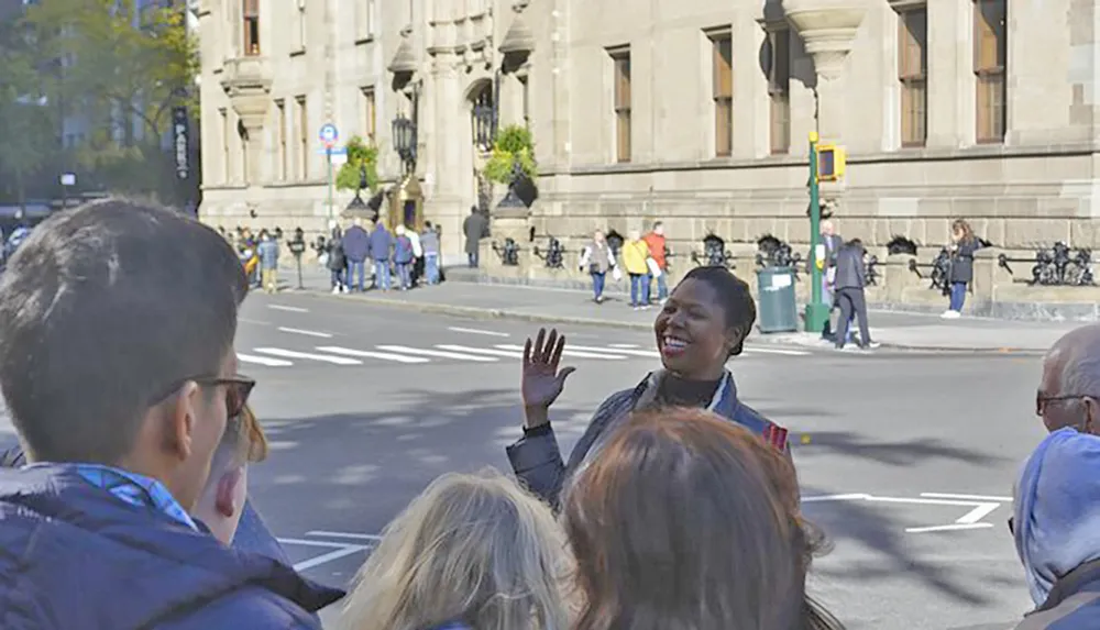 A smiling woman is gesturing and speaking to a group of people on a city street possibly leading a walking tour