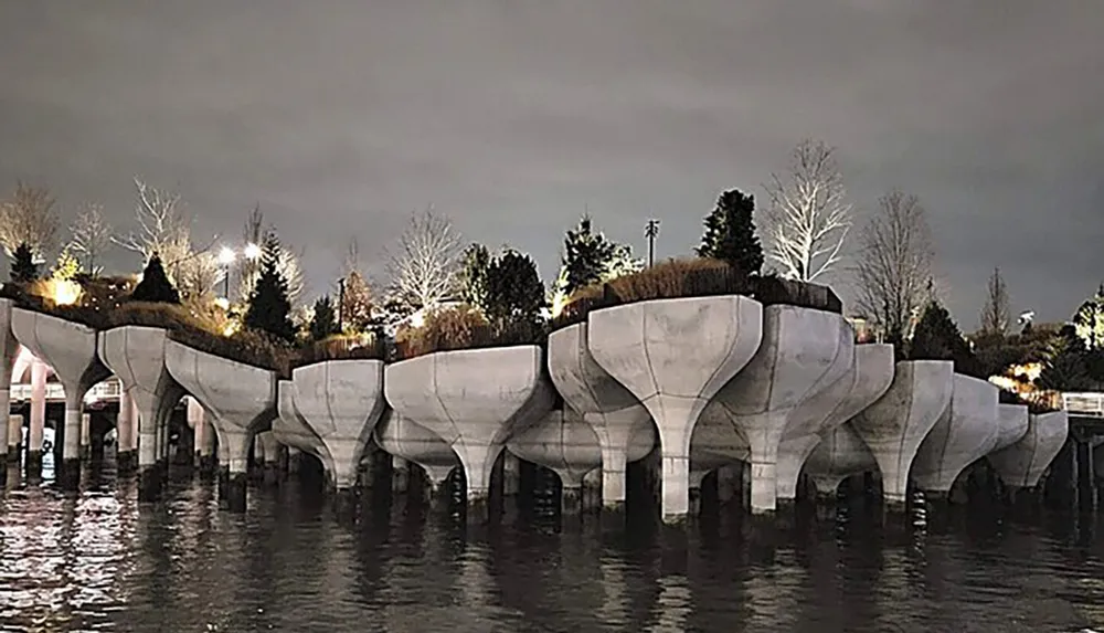 The image shows a series of large concrete flowerpot-shaped structures holding trees and plants standing in water at dusk or night time creating an intriguing and surreal landscape