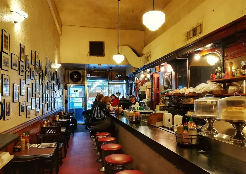 The image depicts the cozy interior of a classic diner with patrons seated at the counter and framed pictures lining the wall