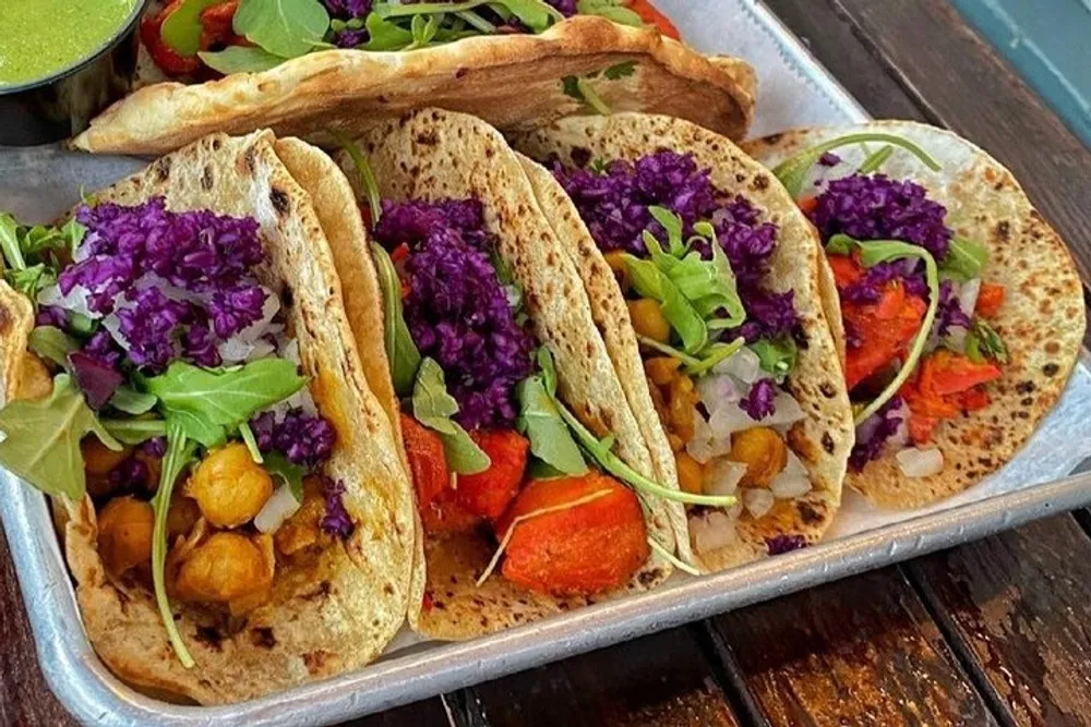 The image shows a tray of vibrant vegetable-filled tacos with a bright purple cabbage garnish and a side of green sauce