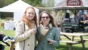 Two smiling women are holding drinks at an outdoor event with tents and a sign reading 