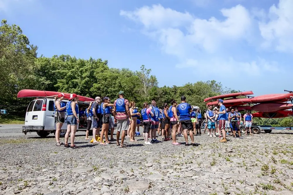 A group of people wearing life jackets and carrying kayaks is preparing for a river adventure under a clear blue sky