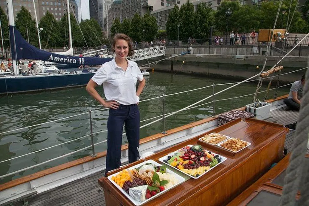 A woman stands smiling on a boat next to a buffet spread of fruits cheeses and other snacks with another boat and a waterfront promenade in the background
