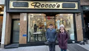 A young man and woman are smiling in front of Pasticceria Rocco, a bakery with a cozy interior visible through its glass facade.