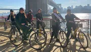 A group of smiling people wearing helmets are standing with bicycles on a sunny boardwalk by the water, with a bridge in the background.
