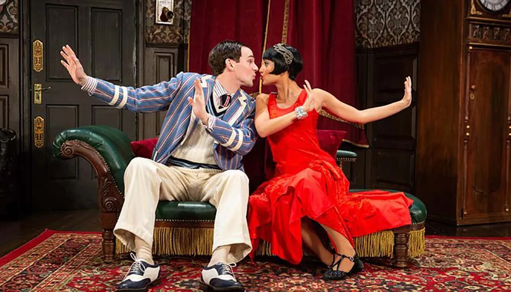 Two actors are performing in a scene on stage where one is seated wearing a striped blazer and reacting with surprise while the other dressed in a red flapper dress leans in as if to kiss him