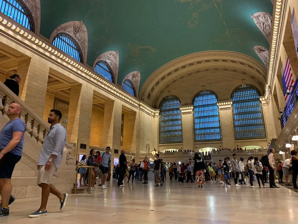 The photo depicts the bustling interior of Grand Central Terminal with its iconic celestial ceiling and grand windows filled with people in motion suggesting a typical busy day at the station