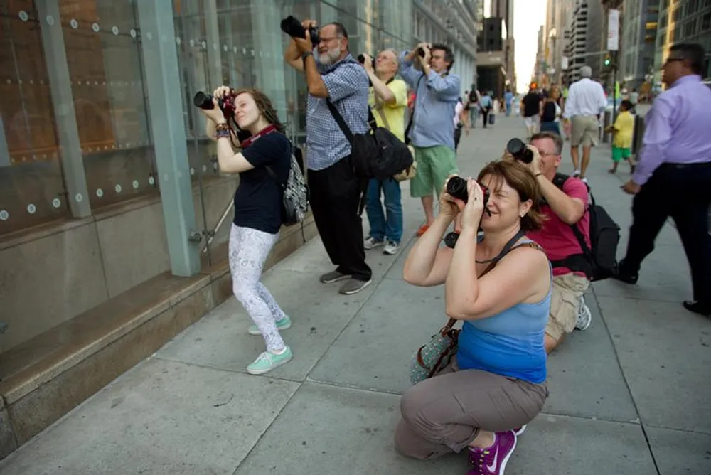 Several individuals are photographing something off-camera on a city sidewalk some using unique stances to capture their shot