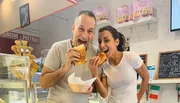 Two people are joyfully biting into large sandwiches inside a bakery or cafe with a cheerful and lively atmosphere.