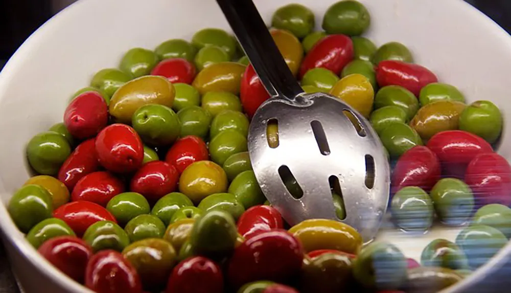 A bowl filled with colorful olives is accompanied by a slotted spoon ready for serving