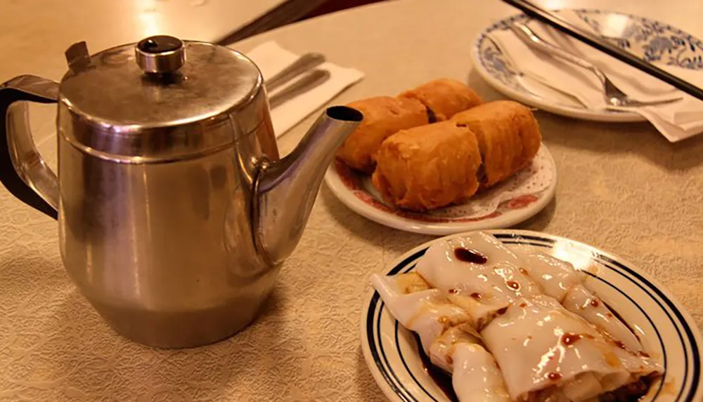 The image features a traditional Chinese tea setting with a metal teapot and dishes containing dim sum items including rice noodle rolls and deep-fried pastries on a table with a patterened tablecloth