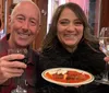 A smiling man and woman are holding glasses of red and white wine respectively with a plate of what appears to be cannoli between them sharing a cheerful moment