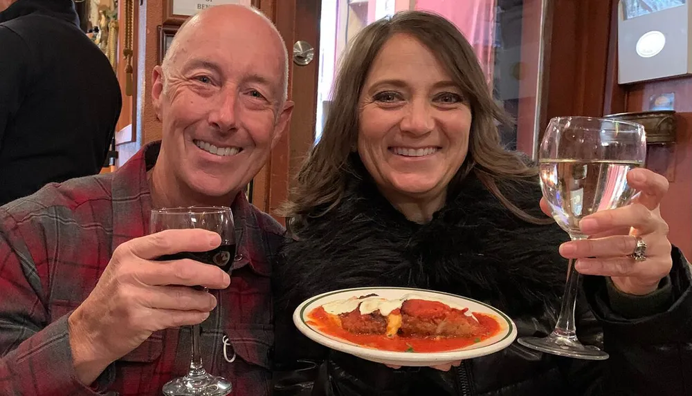 A smiling man and woman are holding glasses of red and white wine respectively with a plate of what appears to be cannoli between them sharing a cheerful moment