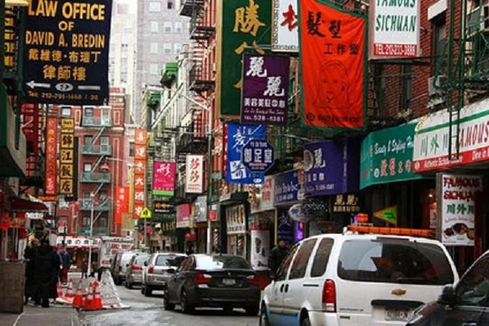 The image shows a bustling street in Chinatown with various signage in Chinese characters cars parked along the street and pedestrians walking