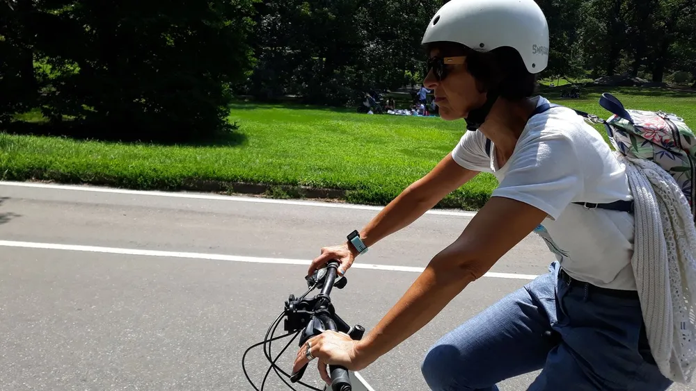 A person wearing a helmet is riding a bicycle on a sunny day with greenery and people in the background