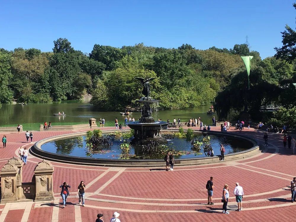 The image shows the bustling Bethesda Terrace and Fountain in Central Park New York with people walking around and enjoying the sunny day by the water