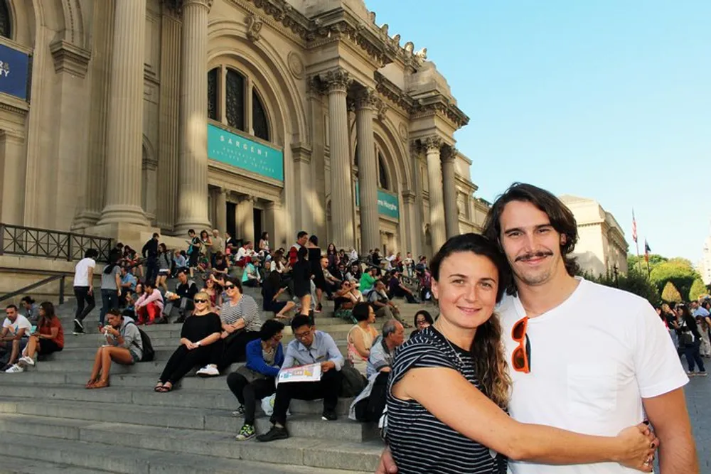 A couple is posing for a photo in front of a bustling set of steps leading up to what appears to be an iconic museum entrance