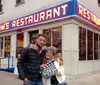 The image shows the exterior of Toms Restaurant with blue umbrellas and chairs outside and banners referencing the television show Seinfeld