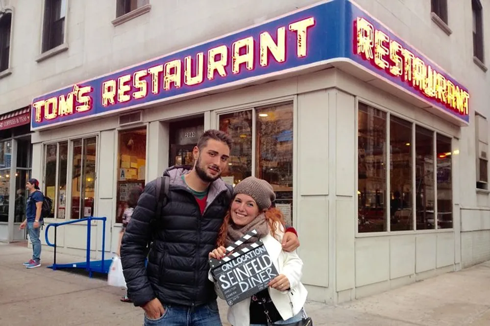 A couple is smiling in front of Toms Restaurant famously known for its exterior being used in the sitcom Seinfeld while the woman holds a sign that reads On Location Seinfeld Diner