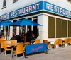 The image shows the exterior of Toms Restaurant with blue umbrellas and chairs outside and banners referencing the television show Seinfeld