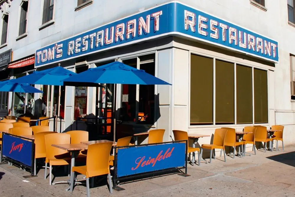 The image shows the exterior of Toms Restaurant with blue umbrellas and chairs outside and banners referencing the television show Seinfeld