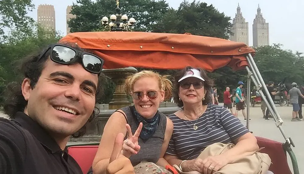 Three people are taking a smiling selfie while seated in a pedicab in a park with city buildings in the background