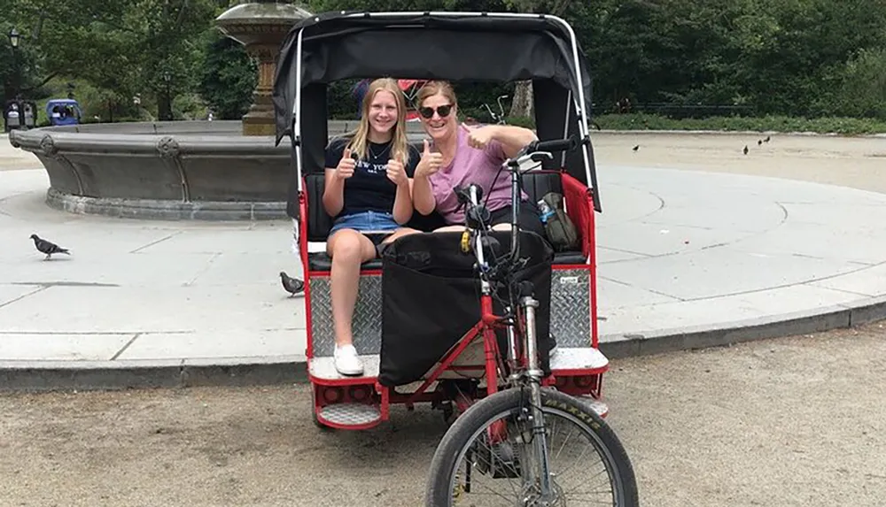 Two people are giving thumbs up while sitting in a pedal-powered rickshaw in a park