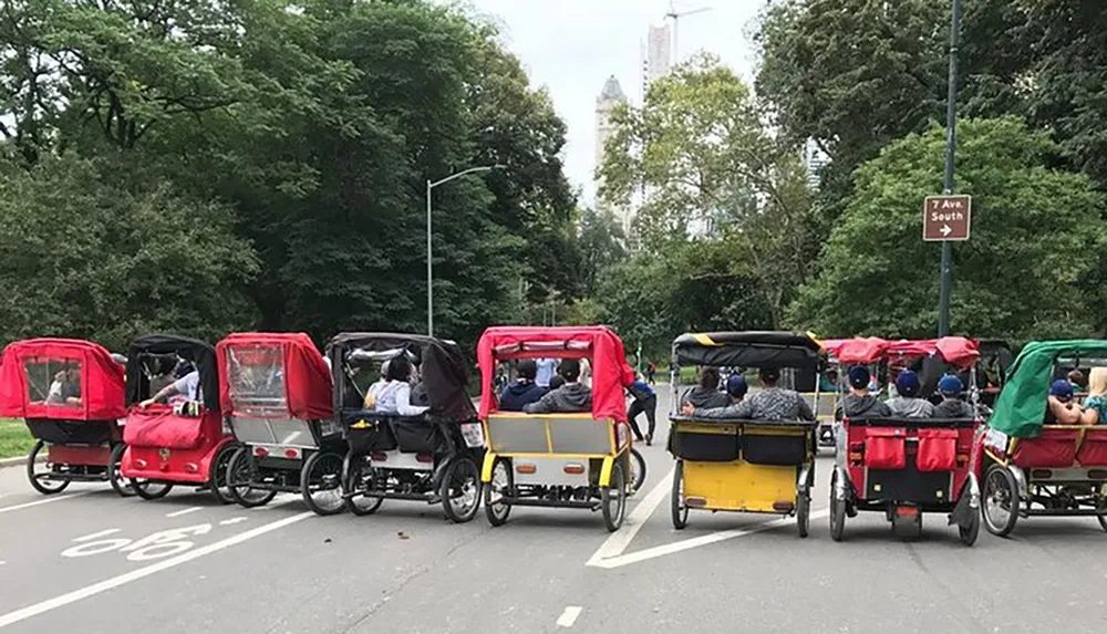 A line of colorful pedicabs transport passengers down a park road with trees and a city skyline in the background