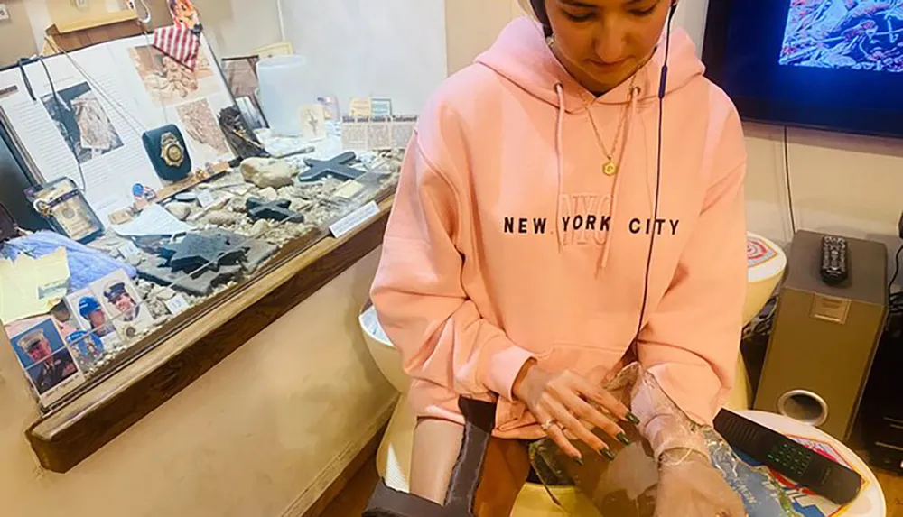 A person in a pink hoodie with NEW YORK CITY written on it sits at a table with a display of various items and memorabilia in the background