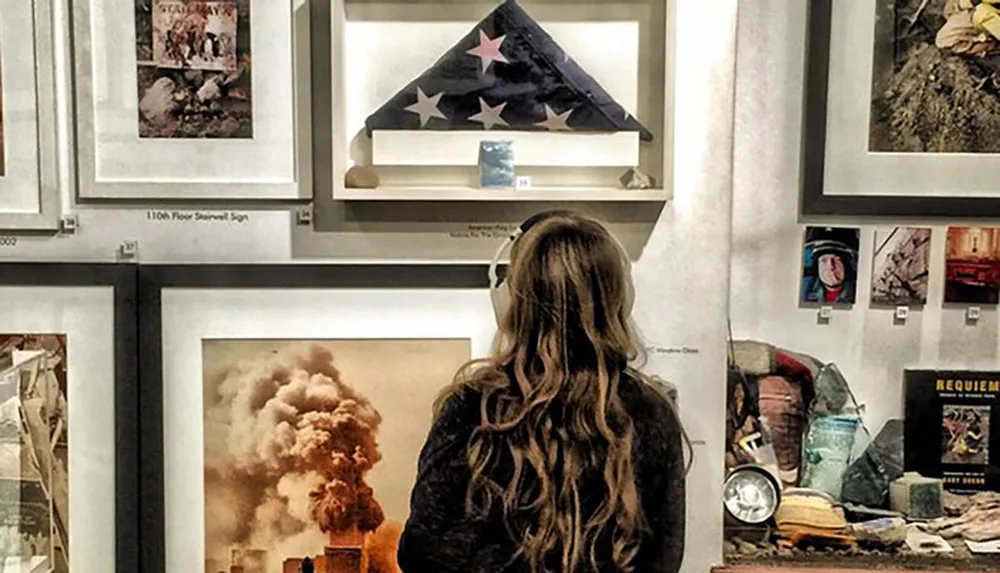 A person is observing a collection of framed photographs and artifacts on a gallery wall which seems to evoke a sense of historical significance or remembrance