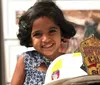 A joyful young child with curly hair and a clip is smiling behind a firefighters helmet with a badge reading Honorary Battalion Chief