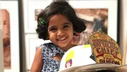 A joyful young child with curly hair and a clip is smiling behind a firefighter's helmet with a badge reading 