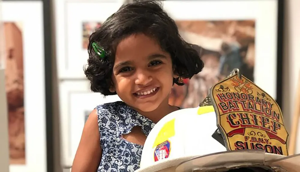 A joyful young child with curly hair and a clip is smiling behind a firefighters helmet with a badge reading Honorary Battalion Chief