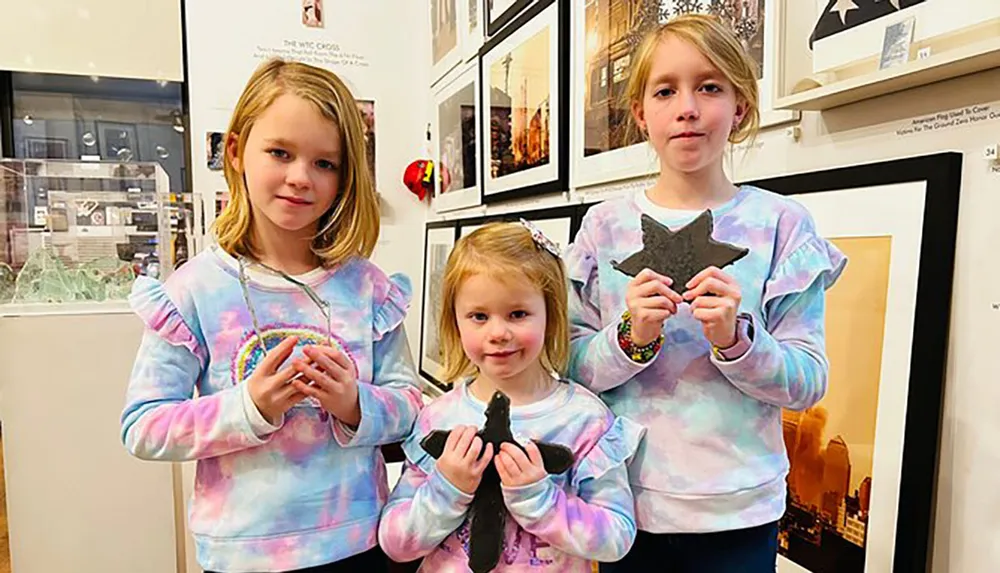 Three children in matching tie-dye sweatshirts are smiling and holding up handmade crafts with framed pictures and exhibits in the background