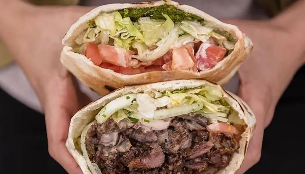 A person is holding a cut open meat-filled pita sandwich revealing the lettuce tomatoes and other fillings inside