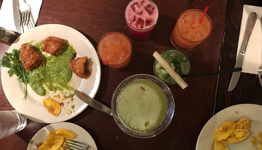 The image shows a dining table from a top-down perspective with multiple dishes including what appears to be fried food with green sauce plantains and several glasses of colorful beverages
