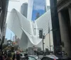 The image shows pedestrians and vehicles on a busy street with the distinctive modern white structure of the Oculus in New York City in the background
