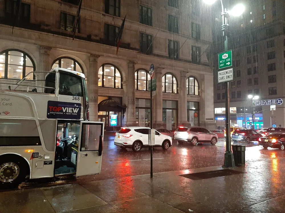 A double-decker sightseeing bus is parked on a rain-soaked street at nighttime with urban traffic and city lights around it