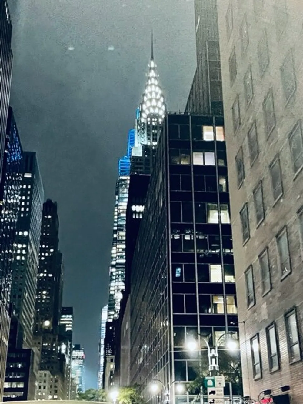 The image captures a nighttime view of a city street flanked by illuminated high-rise buildings with the iconic art-deco spire of the Chrysler Building prominent against a cloudy sky