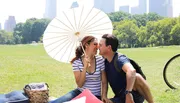 A couple enjoys a sunny picnic in a grassy park with a white parasol and the city skyline in the background.