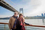 A couple is enjoying a sunny day on a boat cruise with a scenic suspension bridge in the background.