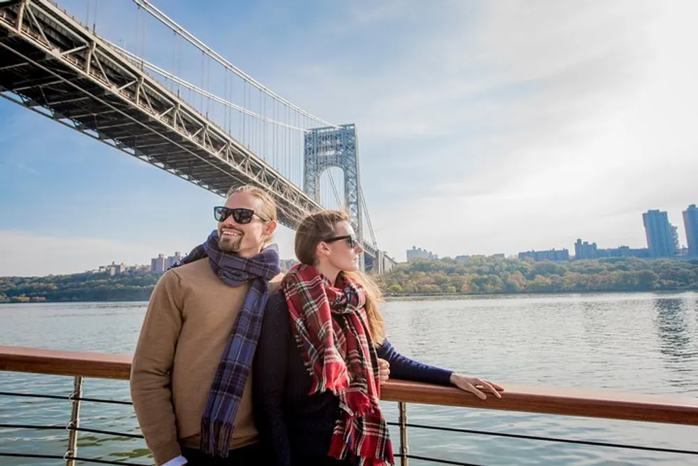 A couple is enjoying a sunny day on a boat cruise with a scenic suspension bridge in the background