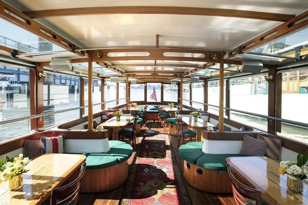 The image shows a cozy and well-appointed interior of a boat with dining tables cushioned bench seating and an elegant wood finish bathed in natural light from surrounding windows